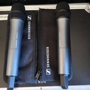 Dvě bezdrátové handheld mikrofony stříbrno-šedé barvy s černými mřížkami. Na těle obou mikrofonů je nápis SENNHEISER XS WIRELESS. Mikrofony leží na č…