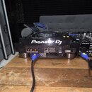 Zadní strana černého DJského přehrávače s nápisem Pioneer DJ a označením modelu CDJ-3000. Jsou vidět různé konektory, do kterých jsou zapojeny modré…