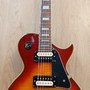Elektrická kytara s tłem v barvě sunburst a zlatým hardwarem. Na hlavě je nápis johnson. Kytara má dva humbuckery, černý plechtrum a čtyři černé ovlá…