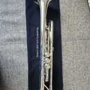 Stříbrná trumpeta položená na tmavě modrém potahu s bílým nápisem TRUMPETS & FLUGELHORNS. Nástroj je v lesklém chromovaném povrchu bez viditelných po…