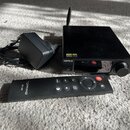 Černý audio přístroj s knoflíkem a anténou, na kterém jsou nápisy TOPPING a DX3Pro. Vedle přístroje je černý napájecí adaptér s kabelem a černý dálko…