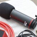 Černý kondenzátorový mikrofon s nasazenou černé pěnovou plopsní. Na těle mikrofonu je bílý nápis Scarlett Studio a červený prstenec u základny. Vedle…