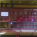 Červený sampler s průhledným plastovým krytem a nápisem electribe sampler KORG. Zařízení má displej, několik otočných knoflíků a mřížku šedých podsví…
