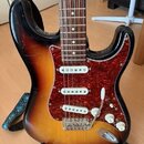 Elektrická kytara s tělem v barvě sunburst a hnědým hmatníkem. Nástroj má červený vzorovaný pickguard a tři bílé snímače. Na těle nejsou viditelné žá…