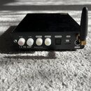Černý obdélníkový elektronický přístroj s připojenou černou anténou. Na zadní straně jsou bílé konektory s nápisy R out, L out, COAX 2, COAX 1 a OPTI…