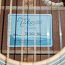 Detail vnitřku akustické kytary s modrou štítkovou nálepkou. Na nálepce je uveden nápis Takamine, G SERIES a Model: GX18CE-NS. Dole je text Crafted i…