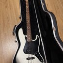 Bílá elektrická basová kytara v černém tvrdém kufru. Na hlavě nástroje je viditelné logo Fender. Kytara má černý pickguard a hnědý hmatník. Povrch ná…