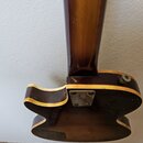 Zadní strana elektrické kytary s tmavě hnědým lakem a světlým lemováním hran. Na kovové spojce krku s tělem je vyryté logo „Gibson“. Povrch lakovanéh…