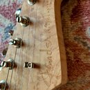 Záběr na zadní stranu hlavy kytary z jasného dřeva s výraznou strukturou slázy. Na dřevě je vyryt nápis Fender STRATOCASTER. Jsou vidět kovové ladičk…