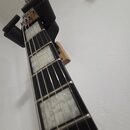 Detail hřednu elektrické kytary s tmavým hmatníkem a velkými bílými vložkami (inlays). Kytara je umístěna v černém nástěnném držáku. Na hlavě nástroj…