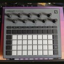 Šedý sekvenční sampler s názvem CIRCUIT RHYTHM a logem Novation v pravém horním rohu. Zařízení obsahuje několik otočných knoflíků, řadu malých tlačít…