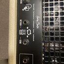 Detail zadní strany kytarového zesilovače s černým povrchem a krémovým potahem. Jsou zde vidět tři zapojovací konektory s popisky REVERB, RETURN a SE…