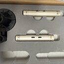 Dvě zlatavé cylindrické kondenzátorové mikrofony umístěné v šedém pěnovém pouzdře. Na jednom z nich je viditelný nápis Made in China a logo CE, na dr…