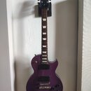 Fialová elektrická kytara s tvarem Les Paul, umístěná na nástěnním držáku. Na hlavě je viditelné zlaté logo Gibson. Kytara má dva humbuckery, čtyři o…
