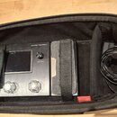 Černý efektový procesor s nápisem AMPERO a označením Amp Modeler / Effects Processor, umístěný v černé polstrované přepravní tašce. Zařízení má displ…