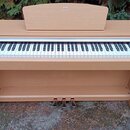 Digitální piano v dřevěné barvě s bílými a černými klávesami. Na horní části je nápis YAMAHA ARIUS. Nástroj má tři pedály a je umístěn venku na beton…