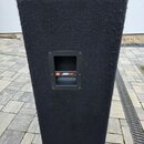 Černý reproduktor s povrchem z texturovaného koberce. Uprostřed je umístěn obdélníkový otvor s nápisem JBL JRX100. Povrch koberce vykazuje mírné opot…