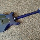 Zadní strana elektrické kytary v tmavě modré barvě s černými kryty elektroniky na těle. Na hridníku je viditelné logo Ibanez. Zadní strana hlavy kyta…