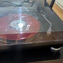 Gramofon s černým základem a průhledným plastovým krytem. Na talíři je položena červená vinylová deska. Nástroj nemá žádné viditelné značky ani nápis…