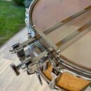 Detailní záběr na horní část snare bubnu s kovovým obručím a mechanismem napnutí ploše. Buben má tělo z dřeva s viditelnou strukturou a kovové části…