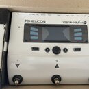 Bílý procesor vokálních efektů s nápisy TC-HELICON a VOICELIVE play GTX. Zařízení má centrální displej s modrými tlačítky označenými jako GENRE, SETU…