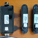 Tři černé elektronické komponenty (pickupy) s bílými nálepkami, položené na dřevěném povrchu. Největší komponenta vlevo má nápis EMG-817H, (2)1289463…