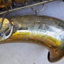 Detail zvonu mosazného saxofonu s viditelným opotřebením povrchu, oxidací a škrábancemi. Na kovu je vyryto logo Selmer a nápis EKHART. V horní části…