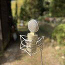 Kondenzátorový mikrofon stříbrné barvy s kovovou mřížkou, umístěný v drátovém shockmountu. Na těle mikrofonu je viditelný nápis „URY“ a pod ním částe…