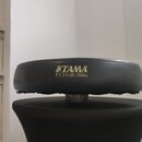 Černý polstrovaný sedák bubeníka s nápisem TAMA a pod ním 1st CHAIR DRUM THRONE SYSTEMS v žluté barvě. Povrch materiálu vykazuje mírné opotřebení a v…