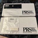 Dvě bílé papírové krabice s černým logem PRS PAUL REED SMITH GUITARS. Na horní krabici je černý papírový pásek s logem PRS GUITARS. Na spodní krabici…