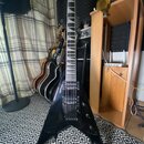 Černá elektrická kytara s tělem ve tvaru V. Na hlavě je viditelné logo Jackson. Kytara má dva humbuckery, fixační most a hриф s vkládanými značkami.…
