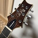 Detail hlavy elektrické kytary s tmavým dřevěným povrchem a bílým lemováním. Na hlavě je bílý nápis SE a stylizované logo Paul Reed Smith. Na hrazenc…