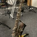 Stříbrná saxofon v pozici na černém stojanu značky HERCULES. Nástroj vykazuje známky používání a povrchové opotřebení. Na těle saxofonu nejsou čiteln…