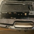 Černý elektronický přístroj v dopasovaném přepravním kufru s pěnovou výplní. Na zadní straně jsou dva kombinované konektory, dva RCA vstupy označené…