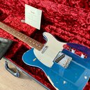 Modrá elektrická kytara s bílým pickguardem v červeném plišovém kufru. Na hlavě kytary je logo Fender a v kufru leží certifikát s nápisem Fender CERT…