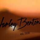 Detailní záběr na povrch nástroje v hnědém odstínu. Je zde vidět černý nápis kurzívou Harley Benton a pod ním menší text est. 1998. Povrch je hladký…