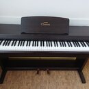 Digitální piano v tmavě hnědé barvě s dřevěnou strukturou. Na notovém pultu je zlatý nápis YAMAHA a Clavinova. Nástroj má standardní klávesnici s bíl…