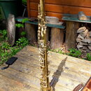 Zlatý mosazný saxofon umístěný na kovovém stojanu. Nástroj je v celkově čistém stavu, bez viditelných značek nebo nápisů. Fotografie je pořízena venk…