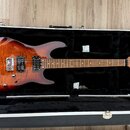 Elektrická kytara s hnědým plamenovitým povrchem těla a hnedým hmatníkem, umístěná v černém tvrdém kufru. Na hlavě kytary je nápis Harley Benton Cust…