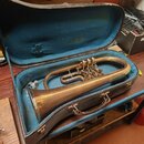 V otevřeném kufru s modrým vnitřkem leží mosazný žiletový nástroj (flugelhorn). Povrch kovu je matný s viditelnými oxidacemi a známky opotřebení. Na…