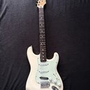 Bílá elektrická kytara typu Stratocaster s jasně zeleným pickguardem. Na hlavě hředu je viditelné logo Fender. Nástroj vykazuje známky používání, jso…