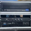 Dvě rackové skříně s audio zařízeními. V horní skříni je černý zesilovač s nápisem PROFESSIONAL AMPLIFIER USA900 a logem QSC. V dolní skříni se nachá…