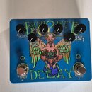 Modrý kytarový efekt s nápisem BAPHOMET a DELAY. Na horní straně jsou čtyři černé otočné knoflíky s popisky REPEATS, TIME (dvakrát) a EFFECT MIX 1. U…