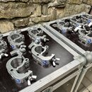 Na dvou černých transportních kufrech z hliníku je rozloženo 14 kovových svorek stříbrné barvy. Každá svorka má na boku nápis SWL 250KGS a modré kula…