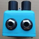 Modrý kytarový efekt v kovovém šasi s dvěma černými oválnými knoflíky nahoře. Na přední straně jsou umístěny dva kovové konektory jack. Povrch je hla…