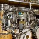Stříbrný kovový snare drum s chromovaným povrchem, umístěný na stojanu. Na korpusu je viditelné logo "Ludwig" v modrém obdélníku a žlutá nálepka s ná…