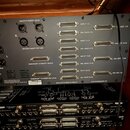 Zadní strana šedého rackového zařízení s nápisy Heritage Audio a MCM-32. Jsou zde vidět různé konektory, včetně několika XLR zdířek a řady D-Sub kone…