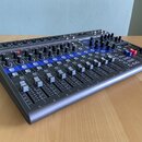 Míchací pult s černým šasi a modrými prvky v horní části. Na ovládacích prvcích jsou viditelné nápisy REC / PLAY, MUTE a COMP. Povrch přístroje vykaz…