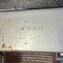 Detail kovového povrchu s nápisem A 00452. Povrch je šedý, v horní části jsou vidět drobné kropky rzi a opotřebení. V horním okraji je vidět část tma…