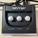 Černý přístroj značky behringer s třemi stříbrnými otočnými ovířkami. Pod ovířkami jsou bílé nápisy MIC/LINE GAIN 1, INST GAIN 2 a OUTPUT. Do horní č…